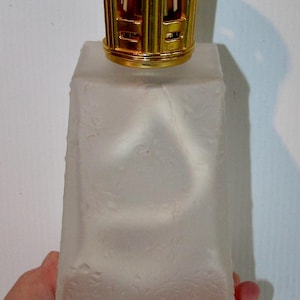 Peut inclure: Une bouteille en verre dépoli avec un dessus en métal doré. La bouteille a une forme rectangulaire avec une surface texturée. Le dessus est un design décoratif en forme de cage.