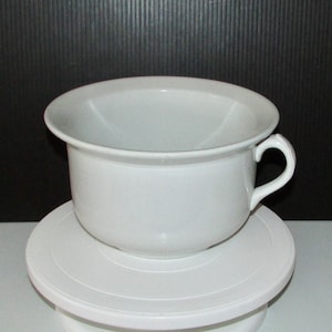 May include: White ceramic chamber pot with a single handle. The pot is sitting on a white cake stand.
