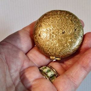 May include: A gold-coloured compact with a floral design. The compact is closed and has a hinged lid.