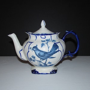 Peut inclure: Une théière blanche avec un motif d'oiseau bleu. La théière a une poignée et un bec verseur bleus. L'oiseau est perché sur une branche avec des feuilles bleues.
