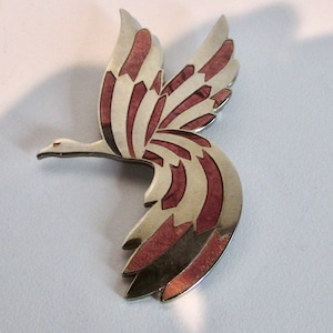 May include: A silver and pink enamel bird brooch with a stylised design. The bird is in flight with its wings spread wide.