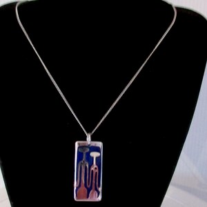 Peut inclure: Collier pendentif argenté avec un pendentif rectangulaire. Le pendentif présente un fond bleu avec deux figures stylisées abstraites en rouge et blanc. Le collier est présenté sur un fond noir.