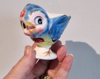 Könnte beinhalten: Ein blauer und gelber Keramik-Eierbecher in Form eines Vogels mit einer roten Blume auf dem Kopf und großen, bemalten Augen. Der Vogel sitzt auf einem weißen Sockel.