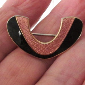 May include: A vintage brooch in a U-shape. It has a gold-tone metal frame, black enamel edges, and a textured copper-coloured centre. A silver-tone pin is fixed to the back. A classic piece of jewellery.