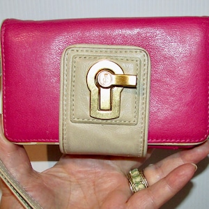 May include: A pink and cream leather wallet with a gold clasp. The wallet has a wrist strap.
