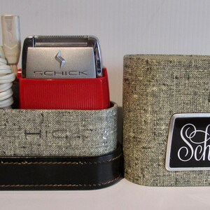 May include: A red and silver electric razor in a grey fabric case with a black leather trim. The case has the word "Schick" printed on it in a silver and black font.