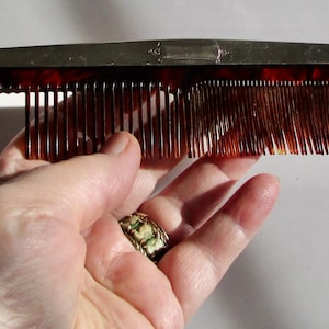 May include: A vintage comb with a tortoiseshell pattern and a silver-tone metal top. The comb has fine teeth and is held in a hand. The comb is a hair care accessory.