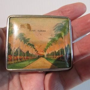 May include: A vintage silver-toned metal compact with a colourful image of palm trees and the text "MIAMI, FLORIDA".