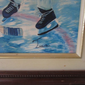 Puede incluir: Una pintura enmarcada de un jugador de hockey patinando sobre hielo. El jugador lleva patines azules y un uniforme blanco. La pintura est&aacute; firmada "Lucie" en la esquina inferior derecha.
