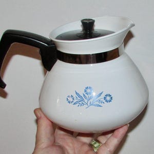 May include: White ceramic coffee pot with a black handle and a silver trim. The pot features a blue floral design.
