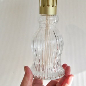 Peut inclure: Un brûleur à huile en verre transparent avec un couvercle en métal doré et une mèche. Le brûleur a un design côtelé et est tenu dans une main.