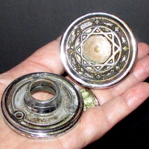 Peut inclure: Deux boutons de meuble vintage en métal argenté. L'un présente un motif d'étoile centrale dans un cadre circulaire, l'autre un centre cylindrique. Les boutons montrent des signes d'âge et d'usure, avec un aspect terni.