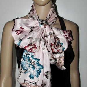 May include: A pink satin scarf with a floral print in shades of teal, burgundy and brown. The scarf is tied in a bow around the neck of a mannequin.