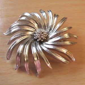 Peut inclure: Une broche argentée en forme de fleur stylisée avec un groupe central de strass.