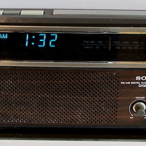 May include: A vintage Sony FM/AM digital clock radio, showing "AM 1:32" in blue digits. The radio has a dark wood grain finish, silver trim, and a speaker grill. The text "SONY FM/AM DIGITAL CLOCK RADIO DIGIMATIC" is visible.