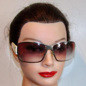 Peut inclure: Lunettes de soleil à monture argentée avec verres dégradés. Les lunettes ont des accents blancs sur les branches. Les verres passent du marron foncé en haut à une teinte plus claire en bas.