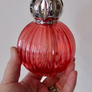 Peut inclure: Lampe à parfum ronde en verre rouge avec un design côtelé et un dessus en métal argenté orné. Le texte "LAMPE BERGER" est visible. La lampe est tenue dans une main.