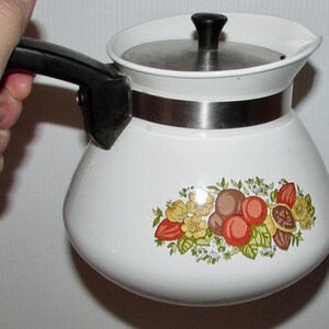 Peut inclure: Cafetière en émail blanc avec une poignée et un couvercle noirs. La cafetière présente un motif floral et fruité décoratif dans les tons de rouge, marron, jaune et vert. Une bande en acier inoxydable entoure le milieu de la cafetière.