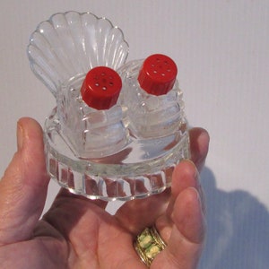 May include: A glass salt and pepper shaker set shaped like a seashell. The shakers have red plastic tops with holes for dispensing salt and pepper.
