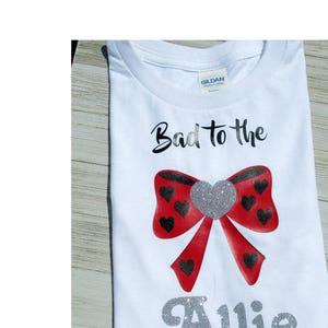 May include: White t-shirt with the text "Bad to the" and a red bow with black hearts. The name "Allie" is written in silver glitter below the bow.