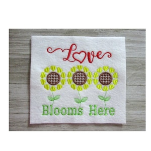 May include: Embroidered white square with three yellow sunflowers, brown centers, and green stems. The word "Love" is in red script above the flowers, and "Blooms Here" is in green script below.