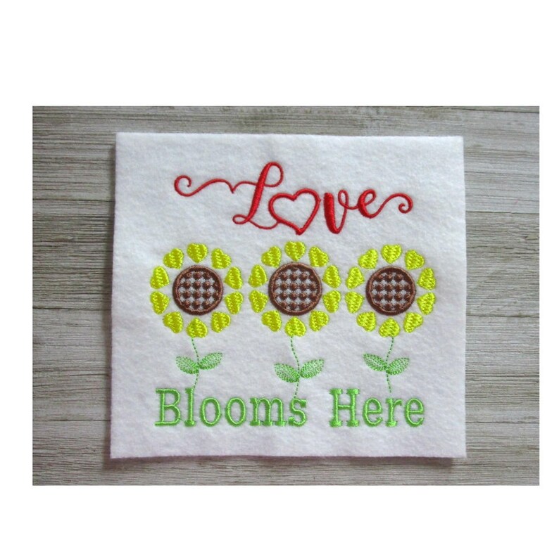 May include: Embroidered white square with three yellow sunflowers, brown centers, and green stems. The word "Love" is in red script above the flowers, and "Blooms Here" is in green script below.