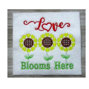 May include: Embroidered white square with three sunflowers and the words "Love Blooms Here" in red and green thread. The sunflowers have yellow petals and brown centers. The text is in a cursive font.