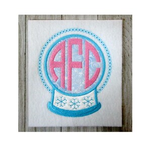 Könnte beinhalten: Ein blauer und rosa bestickter Schneekugel mit den Buchstaben "AFC" im Inneren. Die Schneekugel befindet sich auf einem weißen Hintergrund.