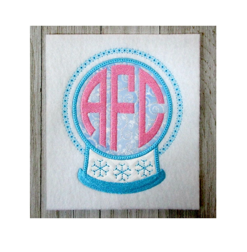 K&ouml;nnte beinhalten: Ein blauer und rosa bestickter Schneekugel mit den Buchstaben "AFC" im Inneren. Die Schneekugel befindet sich auf einem wei&szlig;en Hintergrund.