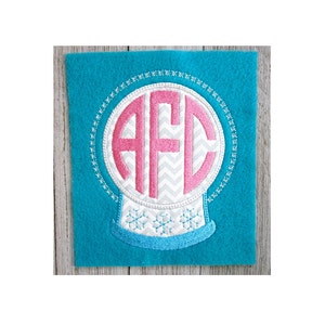 K&ouml;nnte beinhalten: Eine wei&szlig;e Schneekugel mit einem pinkfarbenen Monogramm "AFC" im Inneren. Die Schneekugel ist mit einer blauen Basis und Schneeflocken verziert.