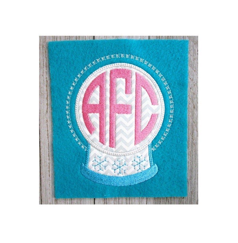 K&ouml;nnte beinhalten: Eine wei&szlig;e Schneekugel mit einem pinkfarbenen Monogramm "AFC" im Inneren. Die Schneekugel ist mit einer blauen Basis und Schneeflocken verziert.
