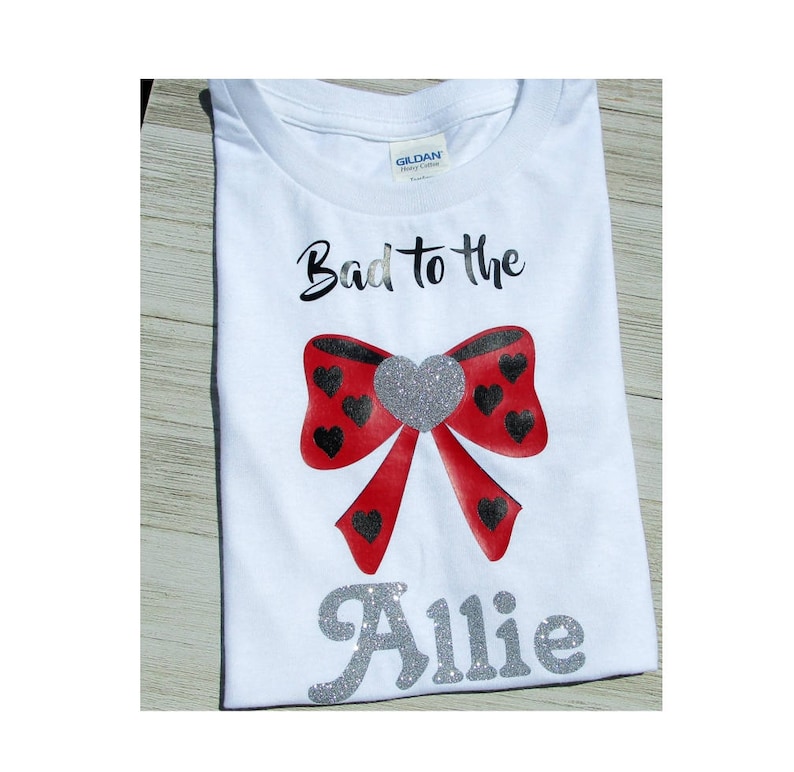 May include: White t-shirt with the text "Bad to the" and a red bow with black hearts. The name "Allie" is written in silver glitter below the bow.