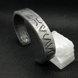 May include: A textured, dark gray metal cuff bracelet with etched markings. The bracelet is open-ended and rests on a white, crystalline rock against a dark background. The bracelet's surface has a unique pattern.
