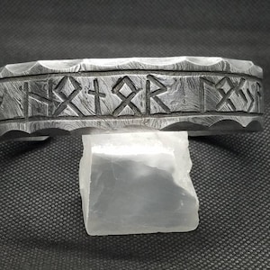 May include: A silver metal bracelet with a hammered finish and engraved runes. The bracelet is on a white surface.