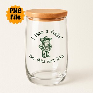 Puede incluir: Un tarro de cristal transparente con tapa de madera. El tarro presenta una ilustraci&oacute;n verde de una rana con sombrero de vaquero y peto. El texto en el tarro dice "I Have a feelin' Your Okies Ain't Dokie."