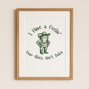 Puede incluir: Una impresi&oacute;n enmarcada con una rana de dibujos animados con sombrero de vaquero y peto. El texto dice "I Have a Feelin' Your Okies Ain't Dokie" en verde, estilo manuscrito. El marco es de color marr&oacute;n claro.
