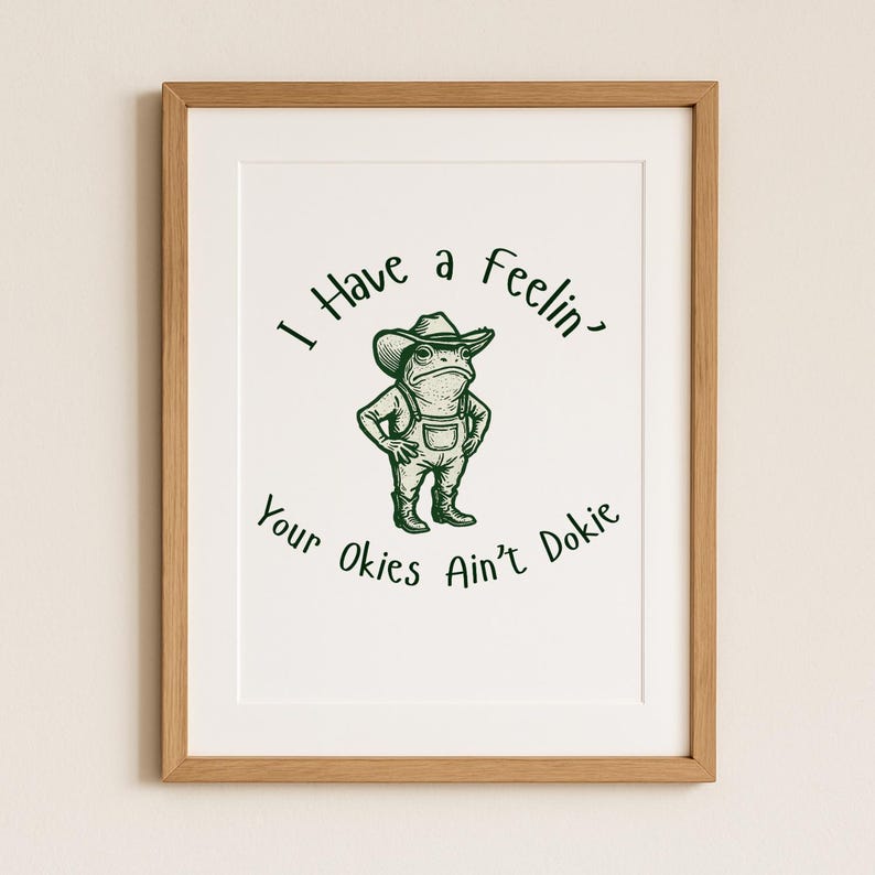 Puede incluir: Una impresi&oacute;n enmarcada con una rana de dibujos animados con sombrero de vaquero y peto. El texto dice "I Have a Feelin' Your Okies Ain't Dokie" en verde, estilo manuscrito. El marco es de color marr&oacute;n claro.