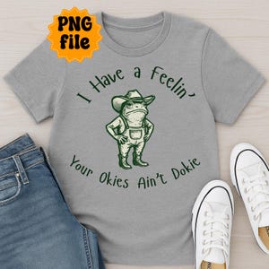 Puede incluir: Camiseta gris con una ilustraci&oacute;n verde de una rana con sombrero de vaquero y peto. El texto "I Have a Feelin' Your Okies Ain't Dokie" est&aacute; impreso encima y debajo del dibujo de la rana. La camiseta se muestra con vaqueros azules y zapatillas blancas.