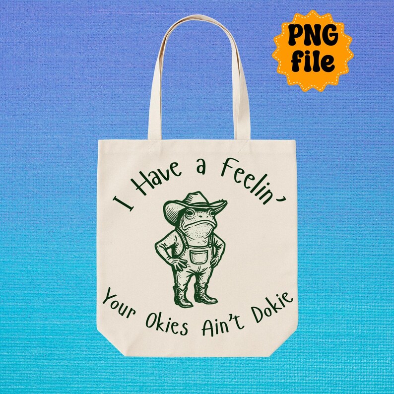 Puede incluir: Bolso tote de color crema con una ilustraci&oacute;n verde de una rana con sombrero de vaquero y peto. El texto verde dice "I Have a Feelin' Your Okies Ain't Dokie." Una pegatina amarilla "PNG file" est&aacute; en la esquina superior derecha.