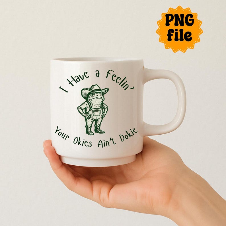 Puede incluir: Taza de cer&aacute;mica blanca con una rana verde de dibujos animados con sombrero de vaquero y peto. La taza tiene el texto "I Have a Feelin' Your Okies Ain't Dokie" en escritura negra. Una pegatina amarilla "PNG file" est&aacute; en el fondo.