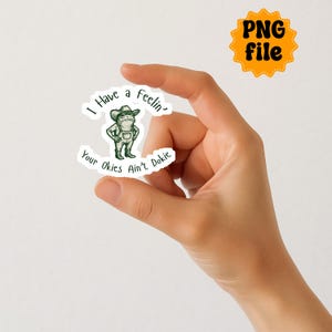Puede incluir: Una pegatina con una rana de dibujos animados con sombrero de vaquero y peto. La pegatina tiene el texto "I Have a Feelin' Your Okies Ain't Dokie" en un dise&ntilde;o curvo. Un icono de archivo PNG naranja separado est&aacute; en la esquina superior derecha.