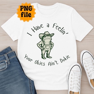 Puede incluir: Camiseta blanca con una ilustraci&oacute;n verde de una rana con sombrero de vaquero y peto. El texto en la camiseta dice "I Have a Feelin' Your Okies Ain't Dokie". Se ven unos vaqueros azules y zapatillas blancas.