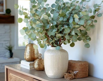 May include: A decorative arrangement featuring a large white vase with vertical lines, filled with eucalyptus branches. Two gold vases and a woven basket sit on a wooden cabinet. The eucalyptus has round, green leaves.
