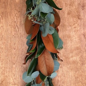 May include: A garland of magnolia leaves and eucalyptus. The garland features large, glossy green leaves and smaller, brown leaves. Sprigs of eucalyptus with round, gray-green leaves are interspersed throughout the garland.