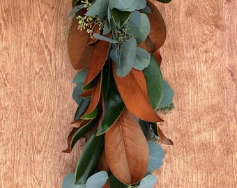 May include: A garland of magnolia leaves and eucalyptus. The garland features large, glossy green leaves and smaller, brown leaves. Sprigs of eucalyptus with round, gray-green leaves are interspersed throughout the garland.