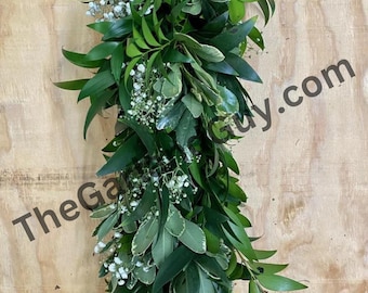 May include: A long garland of green leaves and white baby's breath flowers. The garland is hanging against a wood background.