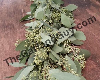 May include: A long garland of eucalyptus leaves and small white flowers. The garland is on a wooden surface.