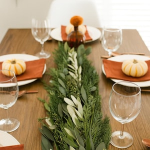 May include: A wooden dining table set for a meal, featuring white plates with orange napkins, mini pumpkins, and clear wine glasses. A green and white garland runs down the center.