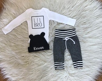 Könnte beinhalten: Ein weißer Langarm-Strampler mit dem Text "Lil Bro" in einem schwarzen Kasten. Eine schwarze Mütze mit dem Namen "Rowan" in weißer Schrift. Grau-schwarze gestreifte Hose mit schwarzem Bund.