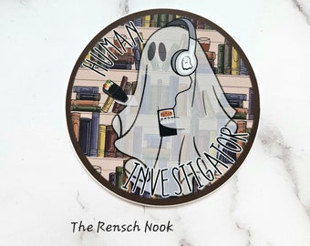 May include: A round sticker with a brown border and a white ghost wearing headphones. The ghost is holding a book and a cup of coffee. The text "Human Investigator" is written around the ghost. The background is a bookshelf with many books.